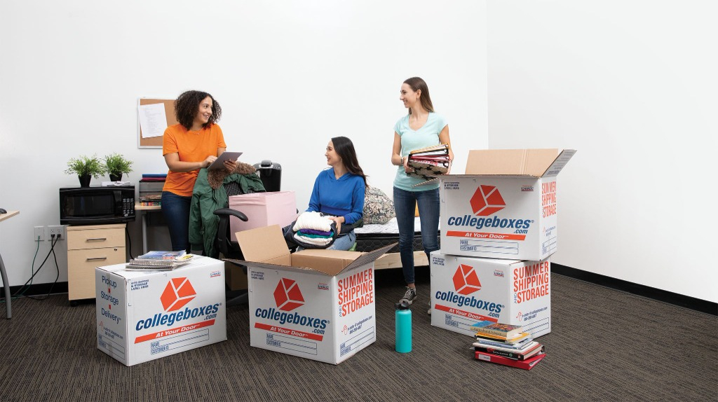 Three women are in a dorm room unpacking, surrounded by boxes.