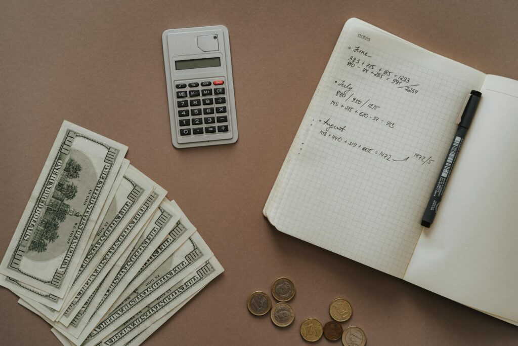 a calculator, notebook, black pen, stack of money on a brown table